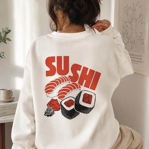 以下が含まれることがあります： 赤い「SUSHI」のグラフィックと寿司のイラストが描かれた白いスウェットシャツ。 デザインには、握り寿司と巻き寿司、さらに漬け生姜とわさびが含まれています。 長袖トップスはカジュアルなスタイルです。