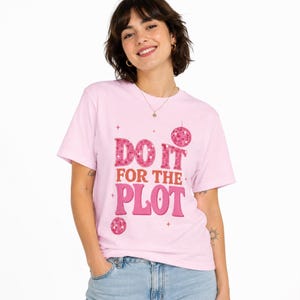 Puede incluir: Camiseta rosa claro con la frase "DO IT FOR THE PLOT" en una tipografía retro. El texto incluye un estampado de cuadros y gráficos de bola de discoteca. La camiseta es de cuello redondo y manga corta.