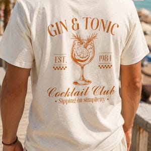 Peut inclure: T-shirt crème avec un motif graphique marron au dos. Le motif présente les mots "GIN & TONIC" au-dessus d'une illustration de verre à cocktail avec le texte "Cocktail Club" et "Sipping on simplicity."