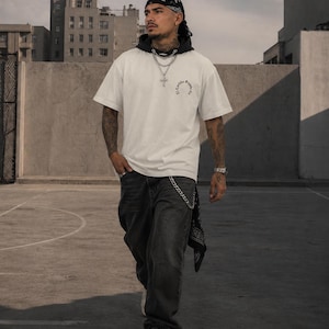 Könnte beinhalten: Eine Person trägt ein weißes T-Shirt mit kleinem Logo, eine schwarze Jeans und weiße Turnschuhe. Die Person trägt auch ein schwarzes Bandana und eine silberne Kette. Der Hintergrund zeigt Stadtgebäude und einen bewölkten Himmel.