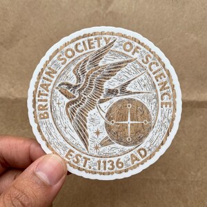 May include: A circular sticker with a wood grain texture featuring a flying swallow, a celestial sphere with a crosshair, and the text BRITAIN SOCIETY OF SCIENCE EST. 1136 AD. The design uses brown and white tones with a scalloped white border edge.