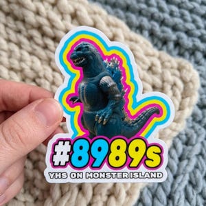 May include: A sticker featuring a blue Godzilla figure with a pink, yellow, and blue outline. The sticker has the text "#8989s VHS ON MONSTER ISLAND" in white and yellow. The sticker is on a textured background.
