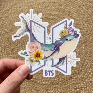 May include: A sticker featuring a watercolor whale design with floral accents, including roses and sunflowers. The whale is set against a purple background with the letters "BTS" at the bottom. The design incorporates sunburst elements.