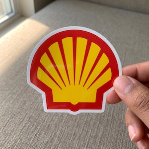 May include: A die-cut sticker shaped like a scallop shell featuring a vibrant yellow center with radiating segments enclosed by a bold red border and a thin white outline, displaying the iconic corporate logo design in a clean graphic style.