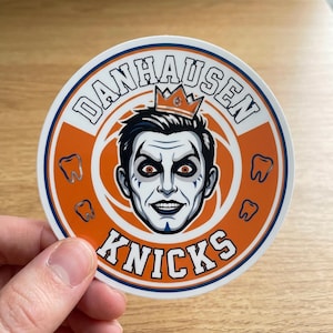 May include: A round sticker with an orange and blue design. The sticker features the text "DANHAUSEN KNICKS" around a central image of a man with white skin, black hair, and a crown. The sticker also has three tooth graphics.