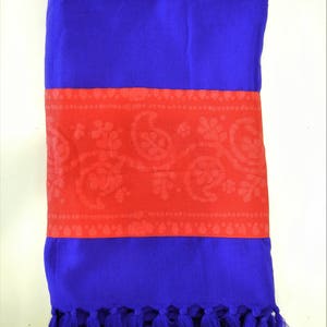 May include: A folded blue scarf with a red band featuring a paisley design. The scarf has a fringe of blue tassels at the bottom. The scarf is made of a soft material.