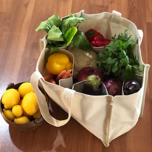 May include: A white canvas tote bag with multiple compartments filled with fresh produce, including yellow peppers, red peppers, cauliflower, purple onions, and green herbs.