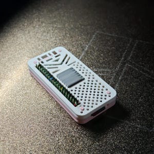 May include: A rectangular electronic device case featuring a white top cover with a perforated grid pattern, a central gray heat sink, and a row of exposed green and gold pins along one side. The base of the case is a soft light pink color.