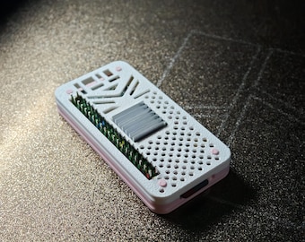 May include: A rectangular electronic device case featuring a white top cover with a perforated grid pattern, a central grey heat sink, and a row of exposed green and gold pins along one side. The base of the case is a soft light pink colour.