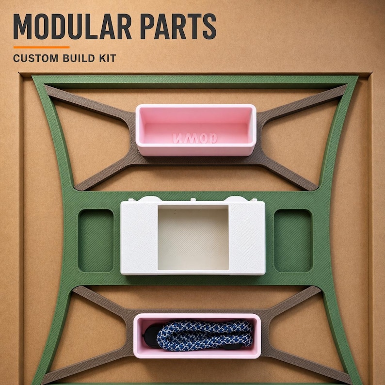 May include: A modular parts custom build kit featuring a green and brown frame with two pink rectangular containers and one white rectangular component. The top pink container has the word MODULAR embossed inside. A blue and white patterned cord is inside the bottom pink container.