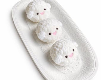 May include: Three small, white, crocheted sheep figures with textured, wool-like surfaces, black bead eyes, and pink stitched mouths. The sheep are arranged in a line on an oval, white, textured ceramic tray with a decorative lace-like pattern.
