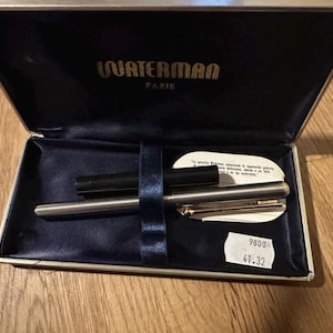 Può includere: Una penna stilografica Waterman Paris argentata in una scatola foderata di velluto blu. La scatola è aperta, rivelando la penna, un cappuccio nero e una piccola etichetta bianca. La scatola è beige con finiture dorate e il logo Waterman.