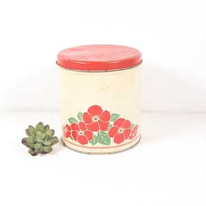May include: A vintage white metal canister with a red lid and a floral design. The canister has a red and green floral pattern with red flowers and green leaves.