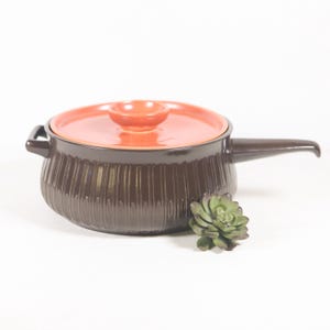 May include: A brown ceramic pot with a ribbed design and a bright orange lid. The pot has a single handle and a small knob on the lid.
