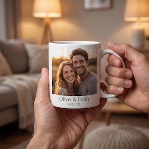 Könnte beinhalten: Ein weißer Keramikbecher mit einem aufgedruckten Foto eines lächelnden Paares. Unter dem Bild steht der Text Oliver & Emily, gefolgt von dem Datum 14.02.2023 in einer dunklen Serifenschrift. Der Becher hat einen Standardhenkel und eine glatte Oberfläche.