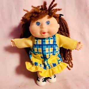May include: A doll with brown yarn hair, blue eyes, and a light skin tone. The doll wears a yellow long-sleeved shirt under a blue and yellow plaid dress with two yellow pockets and a ruffled hem. The doll has white and black shoes on its feet.
