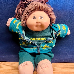 May include: A vintage doll with a soft, vinyl face, brown yarn hair, and a teal outfit. The outfit includes a jacket with the word "OREGON" and a matching shirt and pants. The doll has a seated pose and is on a wooden surface.
