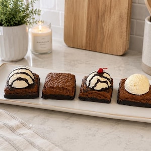 May include: A rectangular white ceramic serving platter holding four square chocolate brownies. Two brownies are topped with scoops of vanilla ice cream drizzled with chocolate syrup, one is plain, and one has ice cream with a red cherry on top.