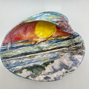 May include: A ceramic seashell-shaped dish with a painted sunset scene. The interior features a yellow sun, red and orange sky, and blue and white ocean waves. The outer edge is a mix of blue, red, and white.