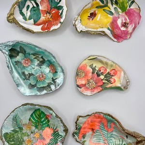 May include: Six decorative oyster shells with floral and tropical designs. The shells feature gold-painted edges and colorful interior artwork, including flowers and a flamingo. The shells are painted with bright colors like pink, orange, and green.