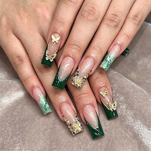 Op de afbeelding: Lange, vierkante kunstnagels met een donker bosgroene French manicure, een glinsterende nude basis en gouden vlinderbedels. Sommige nagels zijn voorzien van gedetailleerde gouden glitteraccenten en kleine doorzichtige strass-steentjes.