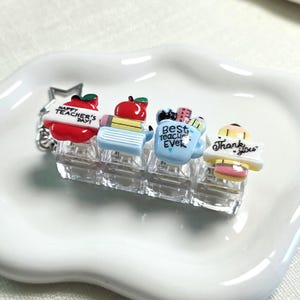 May include: A clear plastic hair clip featuring four decorative charms: a red apple with "Happy Teacher's Day!", a stack of books with a pencil, a light blue mug with "Best Teacher Ever", and a yellow pencil with "Thank You" written in black.