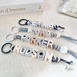 Pode incluir: Quatro porta-chaves personalizados com letras em bloco que formam os nomes EMMA, LUNA, PARKER e HUDSON. Cada porta-chaves inclui um anel de metal, um cordão entrançado e uma mistura de contas com letras prateadas, douradas e creme.