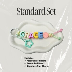 May include: A keychain featuring pastel-colored letter beads spelling GRACE, a pink smiley face bead, and a light green star charm attached by a metal ring. The text Standard Set is above, and a list of included items is below the keychain.