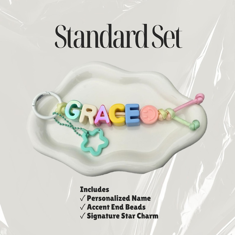 May include: A keychain featuring pastel-colored letter beads spelling GRACE, a pink smiley face bead, and a light green star charm attached by a metal ring. The text Standard Set is above, and a list of included items is below the keychain.
