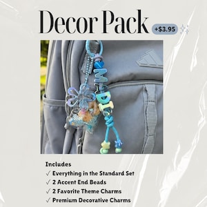 May include: A light blue keychain attached to a gray backpack, featuring the letters MADI in blue beads, a transparent flower charm, a small clear pouch filled with blue and tan beads, and two star-shaped beads at the end of the cord.