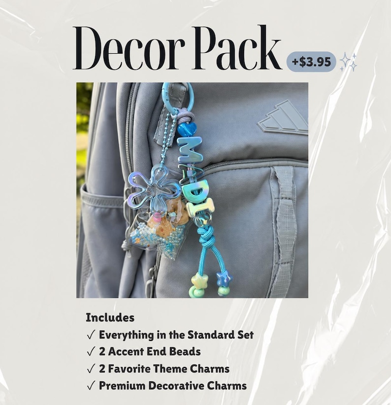 May include: A light blue keychain attached to a gray backpack, featuring the letters MADI in blue beads, a transparent flower charm, a small clear pouch filled with blue and tan beads, and two star-shaped beads at the end of the cord.