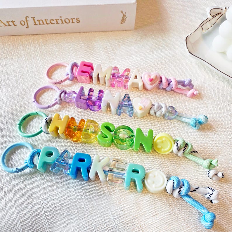 May include: Four colorful keychains featuring plastic letter beads spelling the names EMMA, LUNA, HUDSON, and PARKER. Each keychain includes a circular clasp, translucent and opaque letter beads, heart or smiley face charms, and knotted cord ends.