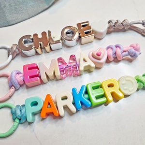Op de afbeelding: Drie gepersonaliseerde sleutelhangers met kleurrijke plastic letterkralen. De bovenste sleutelhanger spelt CHLOE in metallic goud en zilver, de middelste spelt EMMA in roze en crème, en de onderste spelt PARKER in veelkleurige kralen met een smiley.