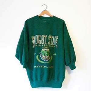 May include: A green Wright State Raiders sweatshirt with the text "WRIGHT STATE RAIDERS" in white and gold lettering. The sweatshirt also features a crest and the text "DAYTON, OHIO". The sweatshirt is hanging on a wooden hanger.