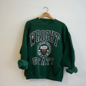 May include: A dark green sweatshirt with the words "WRIGHT STATE UNIVERSITY" in white and black lettering. The sweatshirt has a crew neck and long sleeves, and is hanging on a wooden hanger.