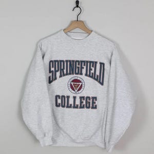 Könnte beinhalten: Ein hellgraues Springfield College Sweatshirt. Das Sweatshirt zeigt die Worte "SPRINGFIELD COLLEGE" in Marineblau und Rot. Ein kreisförmiges Emblem mit einem Dreieck ist unter dem Wort "SPRINGFIELD" zentriert.