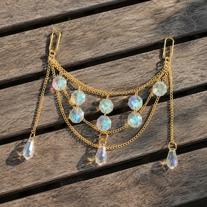 Op de afbeelding: Een goudkleurig kettingaccessoire met iriserende kristallen kralen en druppelvormige hangers. De ketting is gelaagd met meerdere strengen en heeft twee sluitingen. De kristallen reflecteren licht, waardoor een glinsterend effect ontstaat.