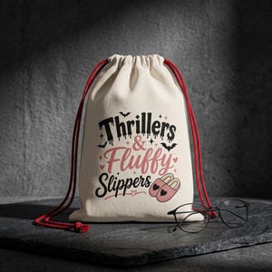 May include: A cream-colored drawstring bag with the text "Thrillers & Fluffy Slippers" in black and pink. The design includes bats, hearts, and slipper illustrations. Red drawstrings and a pair of black-framed glasses are also visible.