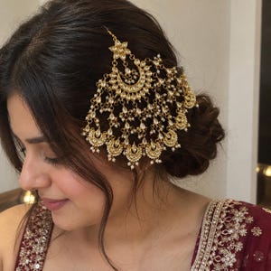 Puede incluir: Un accesorio para el cabello en tono dorado con un diseño grande y complejo en forma de media luna, adornado con piedras transparentes y pequeñas cuentas blancas. La pieza tiene múltiples elementos colgantes que crean un efecto de capas con motivos florales.