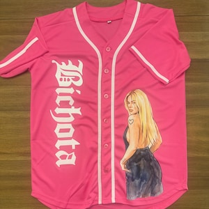 May include: A pink baseball jersey with white accents and the word "Bichota" in white script down the left side. The jersey features an illustration of a woman with long blonde hair and a heart tattoo.