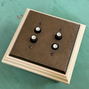 May include: A square light switch plate with a bronze-colored metal faceplate and four white and black knobs. The plate is set in a light-colored wooden frame, with screws visible in the corners.
