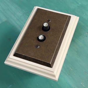 May include: A vintage-style light switch with a brass-colored metal plate and two circular controls. The plate is set within a light-colored wooden frame. The switch has a classic, industrial aesthetic.