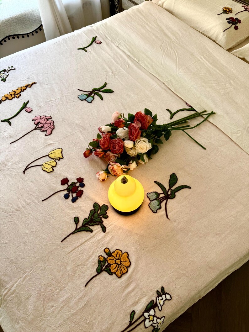 K&ouml;nnte beinhalten: Cremefarbenes Bettw&auml;sche-Set mit gestickten Blumenakzenten. Ein Strau&szlig; Rosen in verschiedenen Farben liegt auf dem Bett, neben einer kleinen, gelben Lampe. Die Bettw&auml;sche umfasst ein Bettlaken, ein Spannbetttuch und Kissenbez&uuml;ge.