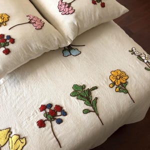K&ouml;nnte beinhalten: Cremefarbenes Bettw&auml;sche-Set mit gestickten Blumenmustern. Das Set umfasst zwei Kissen und einen Bettbezug, verziert mit detaillierten Blumendesigns in Gelb, Rosa, Rot und Blau. Die Blumen sind mit braunen Stielen gestickt.