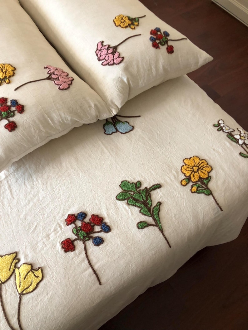 K&ouml;nnte beinhalten: Cremefarbenes Bettw&auml;sche-Set mit gestickten Blumenmustern. Das Set umfasst zwei Kissen und einen Bettbezug, verziert mit detaillierten Blumendesigns in Gelb, Rosa, Rot und Blau. Die Blumen sind mit braunen Stielen gestickt.