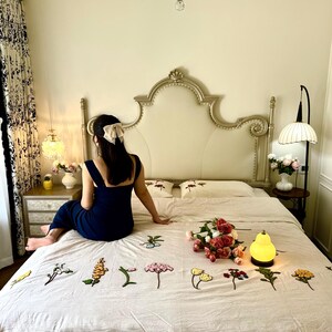 K&ouml;nnte beinhalten: Schlafzimmer-Szene mit einem Bett mit cremefarbenem Kopfteil und floral bestickter Bettw&auml;sche. Ein Strau&szlig; Rosen und eine kleine gelbe Lampe liegen auf dem Bett. Eine Person in einem marineblauen Kleid sitzt auf dem Bett.
