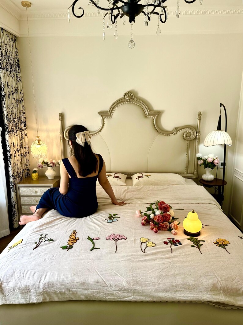 K&ouml;nnte beinhalten: Schlafzimmer-Szene mit einem Bett mit cremefarbenem Kopfteil und floral bestickter Bettw&auml;sche. Ein Strau&szlig; Rosen und eine kleine gelbe Lampe liegen auf dem Bett. Eine Person in einem marineblauen Kleid sitzt auf dem Bett.