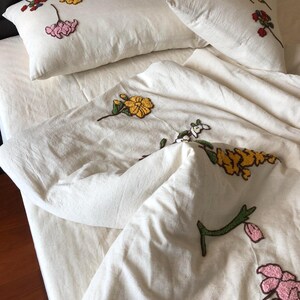 K&ouml;nnte beinhalten: Cremefarbenes Bettw&auml;sche-Set mit gestickten Blumenmustern in Gelb, Rosa und Rot. Das Set umfasst zwei Kissen und einen Bettbezug, alle mit einer strukturierten Oberfl&auml;che. Die Blumenstickerei sorgt f&uuml;r einen Farbtupfer auf dem neutralen Hintergrund.