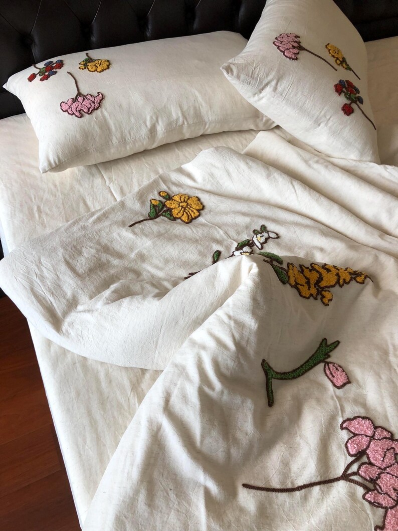 K&ouml;nnte beinhalten: Cremefarbenes Bettw&auml;sche-Set mit gestickten Blumenmustern in Gelb, Rosa und Rot. Das Set umfasst zwei Kissen und einen Bettbezug, alle mit einer strukturierten Oberfl&auml;che. Die Blumenstickerei sorgt f&uuml;r einen Farbtupfer auf dem neutralen Hintergrund.