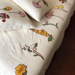 K&ouml;nnte beinhalten: Cremefarbenes Bettw&auml;sche-Set mit gestickten Blumenmustern. Das Set beinhaltet einen Bettbezug und zwei Kissenbez&uuml;ge, verziert mit bunten gestickten Blumen in Gelb, Rosa, Rot und Blau. Die Blumen sind &uuml;ber den Stoff verteilt.